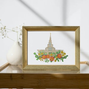May include: A framed print of a watercolor illustration of a white temple with a gold spire surrounded by a wreath of green leaves and orange and yellow flowers.