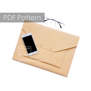 Puede incluir: Una carpeta de estilo sobre de cuero beige con un teléfono inteligente y gafas en la parte superior. La carpeta tiene una solapa que se pliega y se asegura con dos broches metálicos. El texto "LEATHER DIY STUDIO" y "www.leatherdiystudio.com" está impreso en la carpeta.