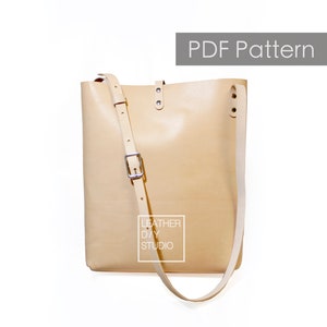 May include: A PDF pattern for a beige leather tote bag with a long adjustable strap. The bag has a simple design with a single pocket on the front. The pattern is for a DIY leather project. "Leather DIY Studio" is printed on the bag.