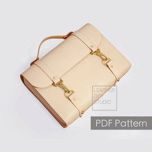 Puede incluir: Un bolso de cuero beige claro con herrajes y hebillas doradas. El bolso tiene un asa superior y una correa ajustable que se puede llevar como bolso de hombro o bandolera. El bolso tiene un cierre de solapa y un pequeño bolsillo en la parte delantera. El bolso está hecho por Leather Day Studio.