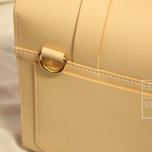 May include: Close-up of a light beige leather bag with a gold metal ring. The bag has a stitched seam and the text "LEATHER D/Y STUDIO" is visible in the lower right corner.