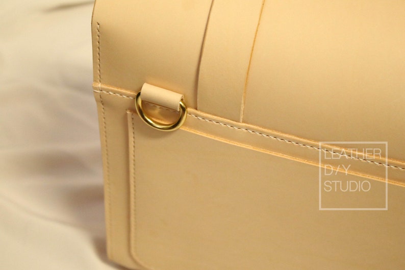 May include: Close-up of a light beige leather bag with a gold metal ring. The bag has a stitched seam and the text "LEATHER D/Y STUDIO" is visible in the lower right corner.
