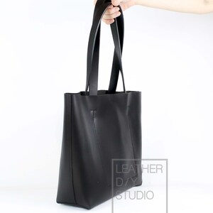 May include: A black leather tote bag with two long straps. The bag is held by a hand and the text "LEATHER DAY STUDIO" is visible on the side.