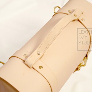 May include: A light beige leather bag with a top handle and brass hardware. The bag has a strap with a buckle and the text "LEATHER D/Y STUDIO" is printed on the side.