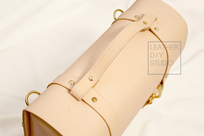 May include: A light beige leather bag with a top handle and brass hardware. The bag has a strap with a buckle and the text "LEATHER D/Y STUDIO" is printed on the side.
