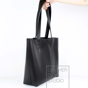 May include: A black leather tote bag with two long straps. The bag is held by a hand and the text "LEATHER DAY STUDIO" is visible on the side.