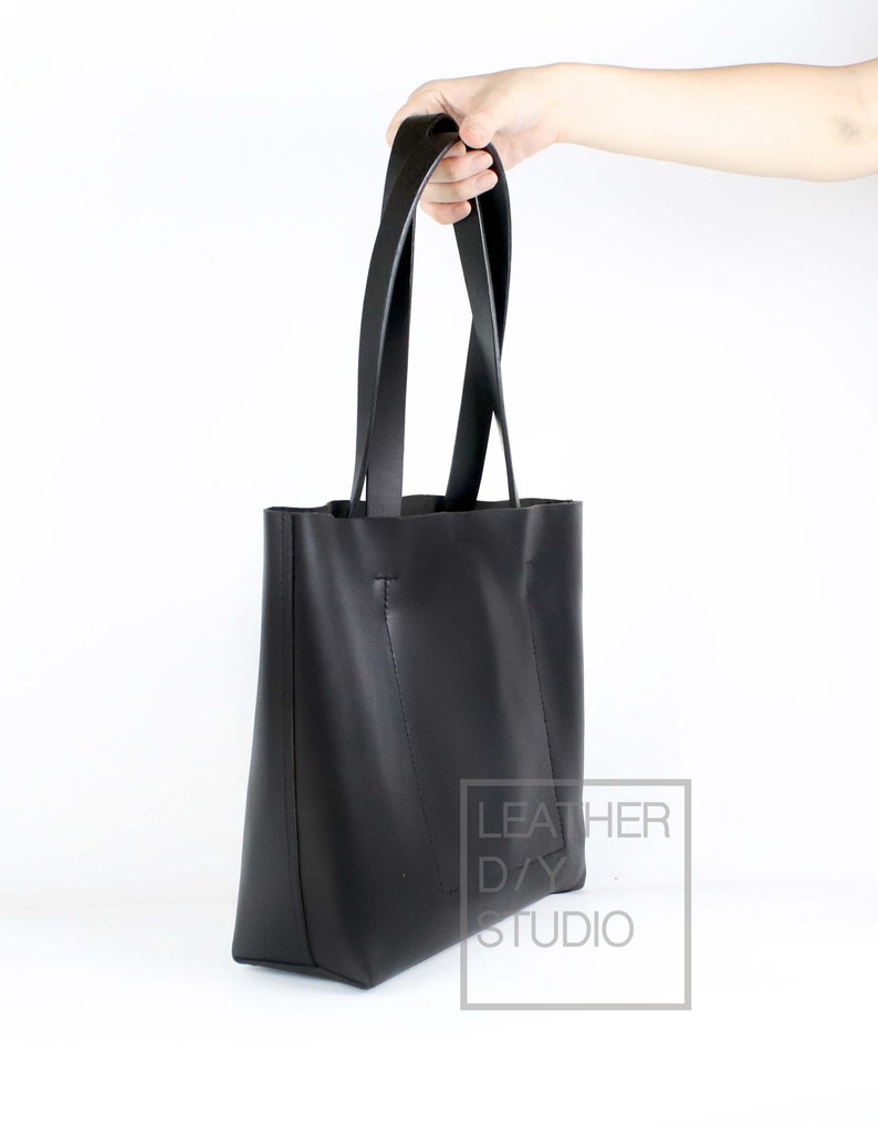 May include: A black leather tote bag with two long straps. The bag is held by a hand and the text "LEATHER DAY STUDIO" is visible on the side.