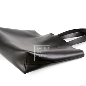 May include: A black leather tote bag with two handles. The bag is lying on its side, with the handles extending out to the right. The bag has a small white label on the side that says "Leather DIY Studio".