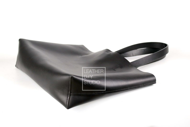 May include: A black leather tote bag with two handles. The bag is lying on its side, with the handles extending out to the right. The bag has a small white label on the side that says "Leather DIY Studio".