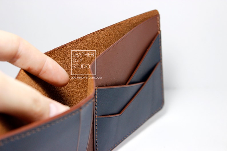 May include: A brown leather wallet with a blue leather interior. The wallet has multiple card slots and a billfold. The wallet is open and the interior is visible. The text "LEATHER D/Y STUDIO" and "LEATHERDIYSTUDIO.COM" is printed on the interior of the wallet.