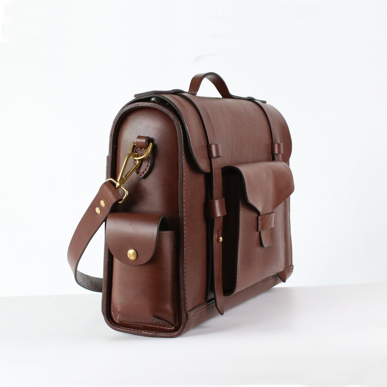 Puede incluir: Un malet&iacute;n de cuero marr&oacute;n con un bolsillo grande en la parte delantera y un bolsillo m&aacute;s peque&ntilde;o en el lateral. La bolsa tiene un asa superior y una correa para el hombro.