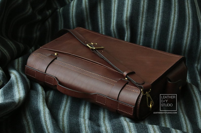 Puede incluir: Un bolso de mensajero de cuero marr&oacute;n con una hebilla de lat&oacute;n y una correa. El bolso tiene una solapa que se cierra con un broche de lat&oacute;n. El bolso est&aacute; sobre una tela a rayas.