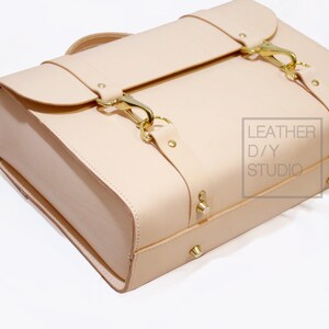 May include: A light beige leather briefcase with gold hardware and buckles. The briefcase has a flap closure and a top handle. The text "LEATHER D/Y STUDIO" is printed on the side of the briefcase.