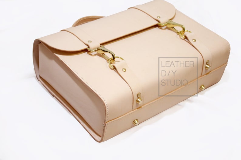 May include: A light beige leather briefcase with gold hardware and buckles. The briefcase has a flap closure and a top handle. The text "LEATHER D/Y STUDIO" is printed on the side of the briefcase.
