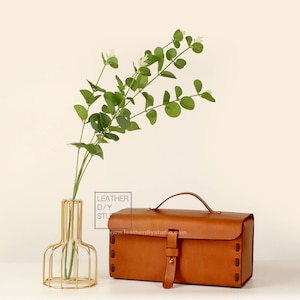 May include: A brown leather box with a brass handle and buckle. The box is sitting on a white surface next to a gold metal vase with a sprig of green leaves.