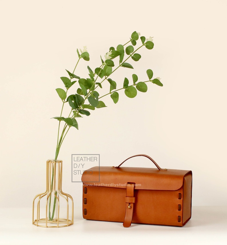 May include: A brown leather box with a brass handle and buckle. The box is sitting on a white surface next to a gold metal vase with a sprig of green leaves.