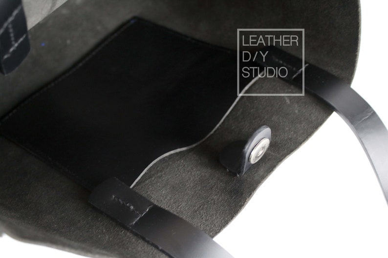 May include: A black leather tote bag with a black leather pocket on the inside. The bag has a snap closure and two black leather handles. The text "LEATHER D/Y STUDIO" is printed on a white rectangle on the bag.