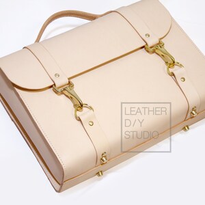 May include: A light beige leather satchel with gold buckles and hardware. The bag has a top handle and a strap. The bag is made by Leather D/Y Studio.
