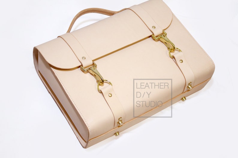 May include: A light beige leather satchel with gold buckles and hardware. The bag has a top handle and a strap. The bag is made by Leather D/Y Studio.