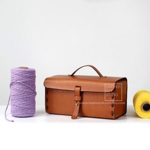May include: A brown leather rectangular bag with a top handle and a buckle closure. The bag has a label that reads "Leather DIY Studio" and a website address. The bag is sitting on a white surface next to two spools of yarn, one purple and one yellow.