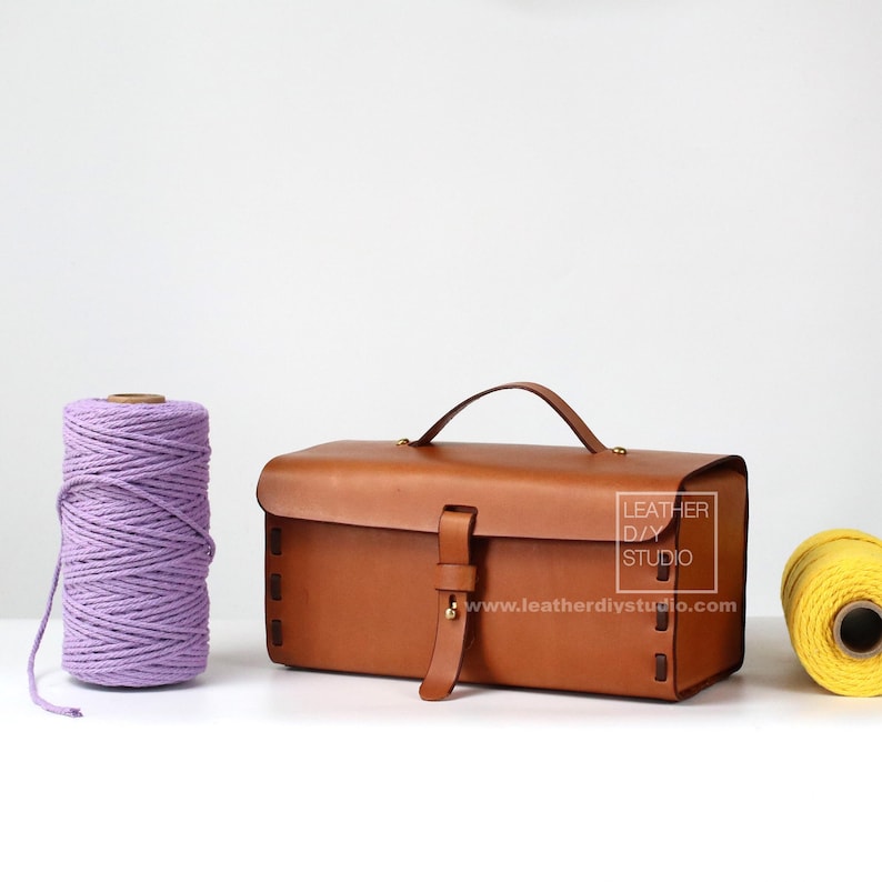 May include: A brown leather rectangular bag with a top handle and a buckle closure. The bag has a label that reads "Leather DIY Studio" and a website address. The bag is sitting on a white surface next to two spools of yarn, one purple and one yellow.