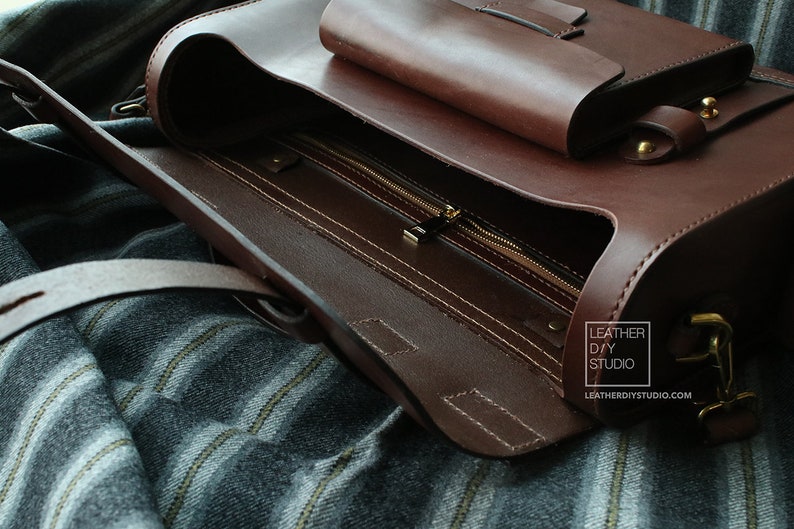Puede incluir: Un bolso de cuero marr&oacute;n con un bolsillo con cremallera y un cierre de solapa. El bolso est&aacute; abierto y el interior es visible. El bolso tiene una correa y una hebilla de metal. El bolso est&aacute; sobre una superficie de tela a rayas. El texto "LEATHER DIY STUDIO" y "LEATHERDIYSTUDIO.COM" es visible en el bolso.