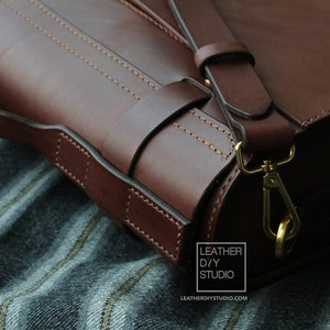 Puede incluir: Primer plano de un bolso de cuero marr&oacute;n con cierre y correa dorados. El bolso tiene un dise&ntilde;o cosido y est&aacute; apoyado sobre una tela a rayas.