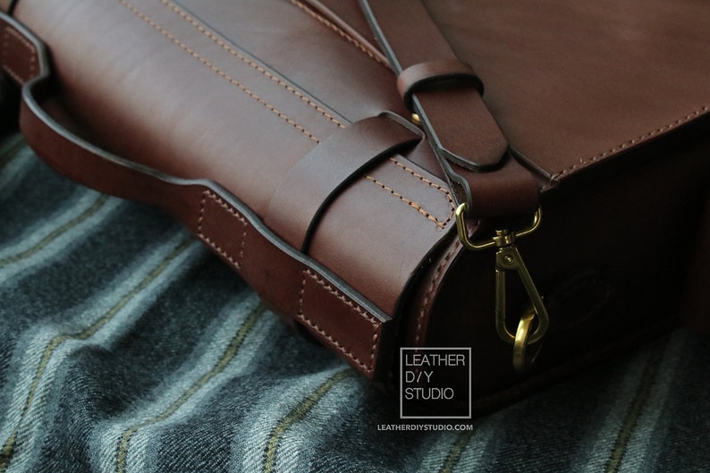 Puede incluir: Primer plano de un bolso de cuero marr&oacute;n con cierre y correa dorados. El bolso tiene un dise&ntilde;o cosido y est&aacute; apoyado sobre una tela a rayas.