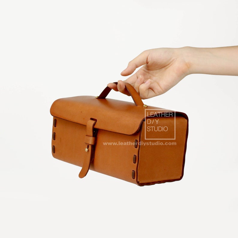 May include: A brown leather satchel bag with a top handle and a buckle closure. The bag has a stitched design around the edges and a small brass label that reads "LEATHER D/Y STUDIO" and "www.leatherdiystudio.com".