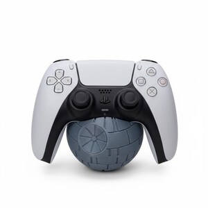May include: A white and black PlayStation 5 controller resting on a gray Death Star-shaped stand. The controller has black joysticks and action buttons. The stand is a detailed replica of the iconic Star Wars space station.