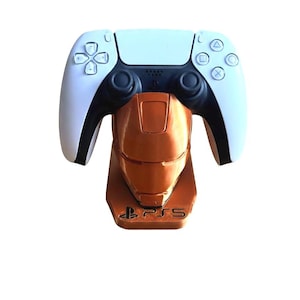 May include: A white and black PlayStation 5 controller rests on an orange stand shaped like a superhero's helmet. The stand has the PS5 logo at the base. The controller has the standard button layout.