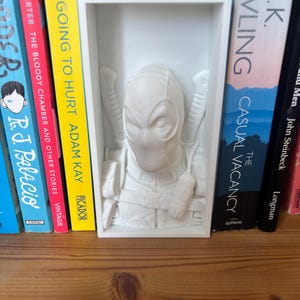 May include: A white Deadpool bust, 3D-printed and framed. The bust shows the character's mask, swords, and utility belt. The frame is on a wooden shelf, with books of various colours behind it. The frame measures approximately 15 cm in height.