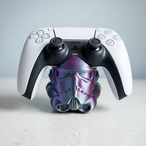 May include: A white and black PlayStation 5 controller resting on a color-shifting, iridescent Stormtrooper helmet-shaped stand. The stand has purple, blue, and silver hues. The controller has black joysticks and buttons.
