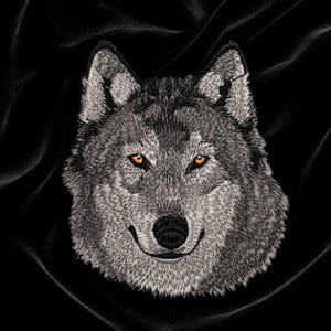 May include: An embroidered patch featuring the detailed head of a gray wolf with textured fur in shades of silver, charcoal, and white. The wolf has piercing amber-colored eyes and a prominent black nose, rendered with intricate stitching detail.
