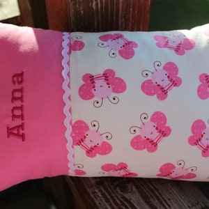 Könnte beinhalten: Ein rechteckiges rosa Kissen mit dem Namen "Anna" in dunklerer rosa Schrift gestickt. Das Kissen hat einen weißen Stoff mit einem Muster aus rosa Schmetterlingen. Eine weiße Spitzenborte trennt die beiden Stoffe.