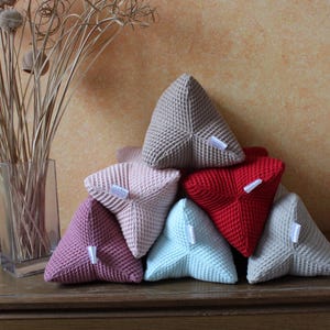 Könnte beinhalten: Ein Set aus sechs dreieckigen Kissen in verschiedenen Farben, darunter Beige, Rosa, Rot und Blau. Die Kissen sind aus einem strukturierten Stoff gefertigt und haben ein schlichtes Design.