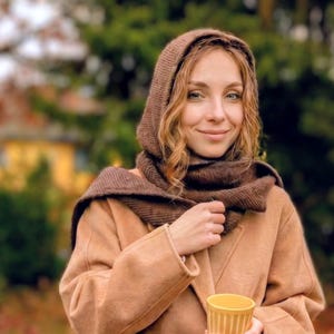 Op de afbeelding: Een vrouw draagt een beige jas met bijpassende sjaal en capuchon, en houdt een gele beker vast. De jas is in de taille dichtgeknoopt. De achtergrond toont bomen en herfstbladeren, wat een herfsttafereel creëert.