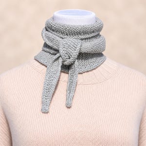 May include: A light gray knitted neck warmer featuring a textured garter stitch pattern. The accessory is wrapped around the neck and secured with a simple front knot, with two pointed ends hanging down over a pale pink ribbed knit sweater.