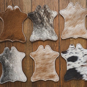 May include: Eight cowhide coasters in various colours and patterns, including brown, grey, and black, arranged on a wooden surface. Each coaster has a unique natural hide pattern and a stitched border.