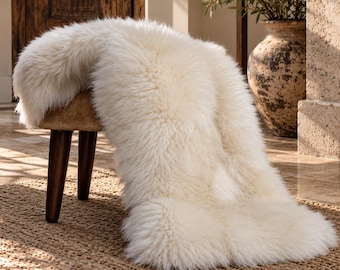 May include: A small stool with dark brown wooden legs and a tan seat, draped with a long, thick, off-white sheepskin rug. The rug features a dense, shaggy texture that covers the top of the stool and extends down toward the floor.