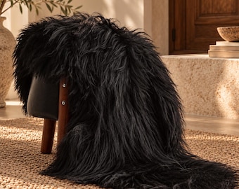 May include: A small stool with three dark brown wooden legs and a black leather seat, draped with a long, thick, black faux fur throw. The fur has a shaggy, textured appearance and hangs down over the side of the stool onto the floor.