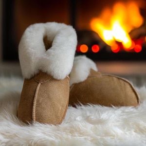 May include: A pair of brown sheepskin slippers with white fur lining. The slippers are sitting on a white fluffy rug in front of a fireplace.