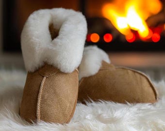 May include: A pair of tan suede slippers featuring a thick, white, plush sheepskin lining that extends over the top edge. The slippers have a rounded toe design with visible stitching along the center seam and a soft, textured exterior finish.