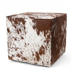 以下が含まれることがあります： 茶色と白の斑点模様の牛革で張られた正方形のオットマンです。