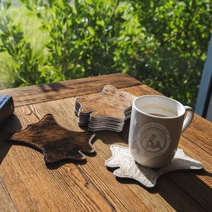 May include: A white coffee mug with a brown logo on a white cowhide coaster. The mug is sitting on a wooden table with a stack of books and another cowhide coaster.