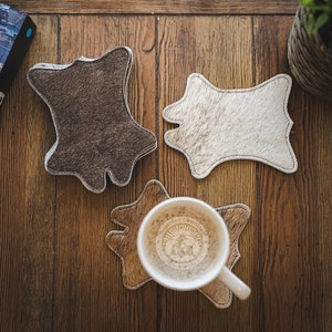 May include: Three cowhide coasters in brown, white, and brown colors. The coasters are in the shape of a cowhide and have a stitched edge. A white coffee mug with a logo is on top of one of the coasters.