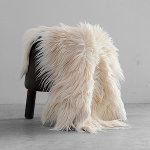 May include: A dark brown stool with three cylindrical legs is draped with a long, shaggy, off-white faux fur throw. The fur has a thick, textured appearance and hangs over the sides of the stool, covering most of the dark seat and legs.