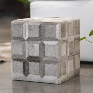 May include: A square gray and white cowhide ottoman with a geometric pattern. The ottoman is sitting on a gray floor.  The ottoman is made of genuine cowhide and is a great addition to any home.