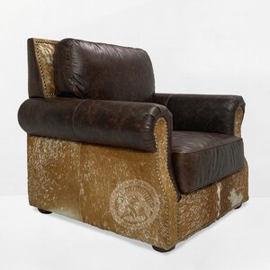 May include: A brown leather armchair with a cowhide base. The chair has a tufted back and a rolled armrest. The chair is accented with brass nailhead trim.