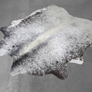 May include: A natural cowhide rug featuring an irregular, organic shape with a speckled pattern of black and dark brown spots against a white background, characterized by a dense, short-haired texture and a distinct central white stripe.